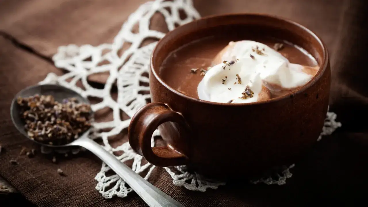 Una tazza di cioccolata calda fumante con panna montata e lavanda, accanto a un cucchiaio pieno di lavanda.