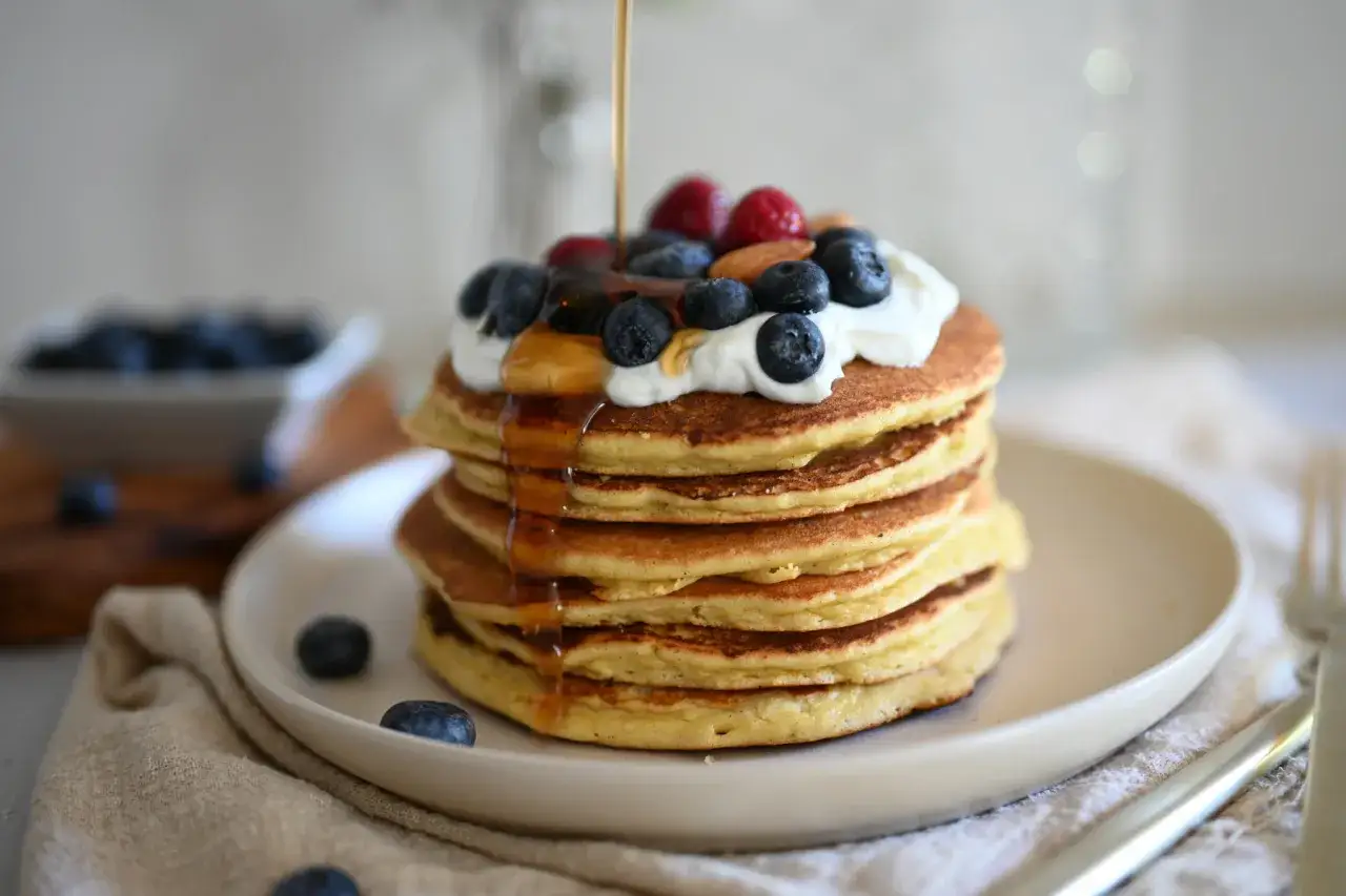 Pancake con yogurt, mirtilli, lamponi e sciroppo d'acero che cola. Una delizia per iniziare la giornata.