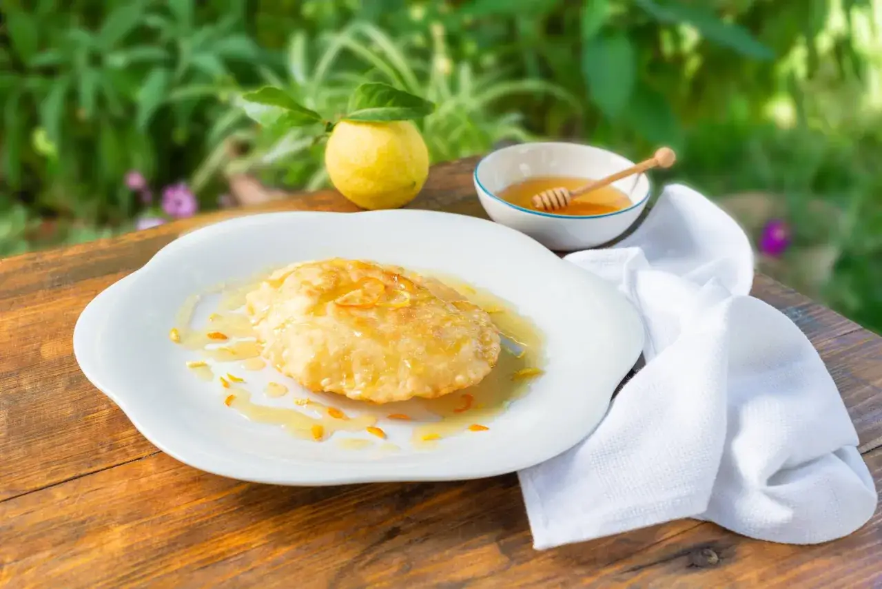 Seadas ricetta: dolce fritto su piatto bianco, irrorato di miele e scorza d'arancia, con limone e miele sullo sfondo.
