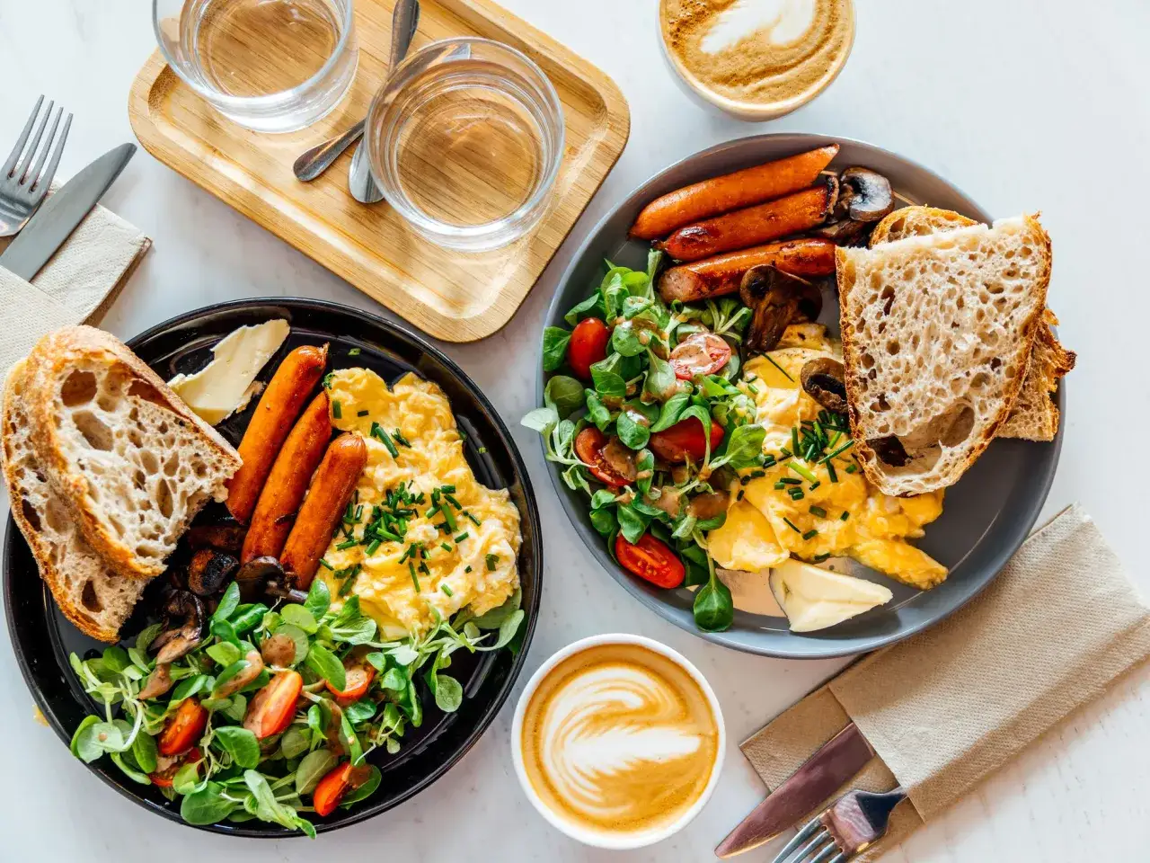 Due piatti di colazione salata con uova strapazzate, salsicce, pane tostato, insalata e caff&egrave;.
