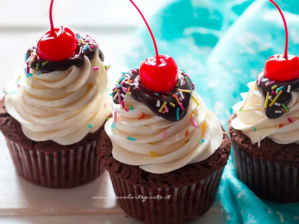 Tre deliziosi cupcake al cioccolato con glassa bianca, scaglie di cioccolato, zuccherini colorati e una ciliegina rossa in cima.