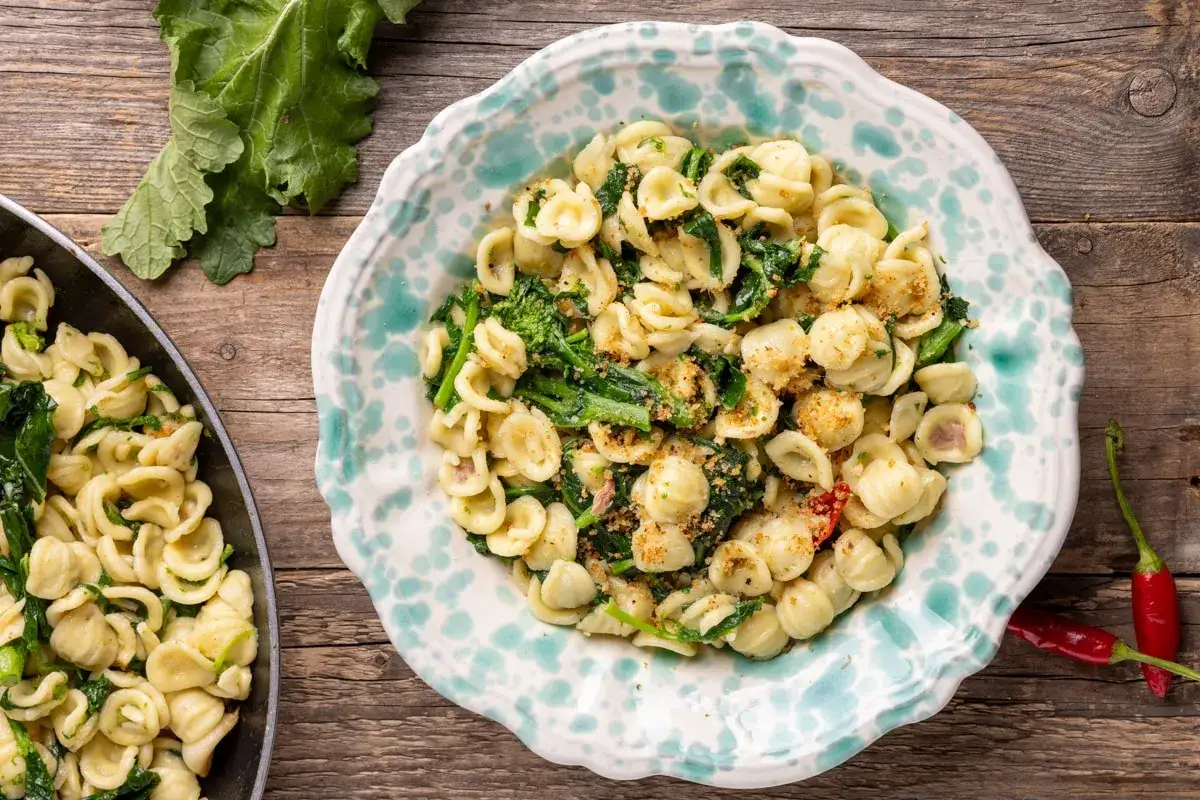 Un piatto di orecchiette cime di rapa fumante, con un contorno di peperoncini rossi e foglie verdi fresche su un tavolo di legno.