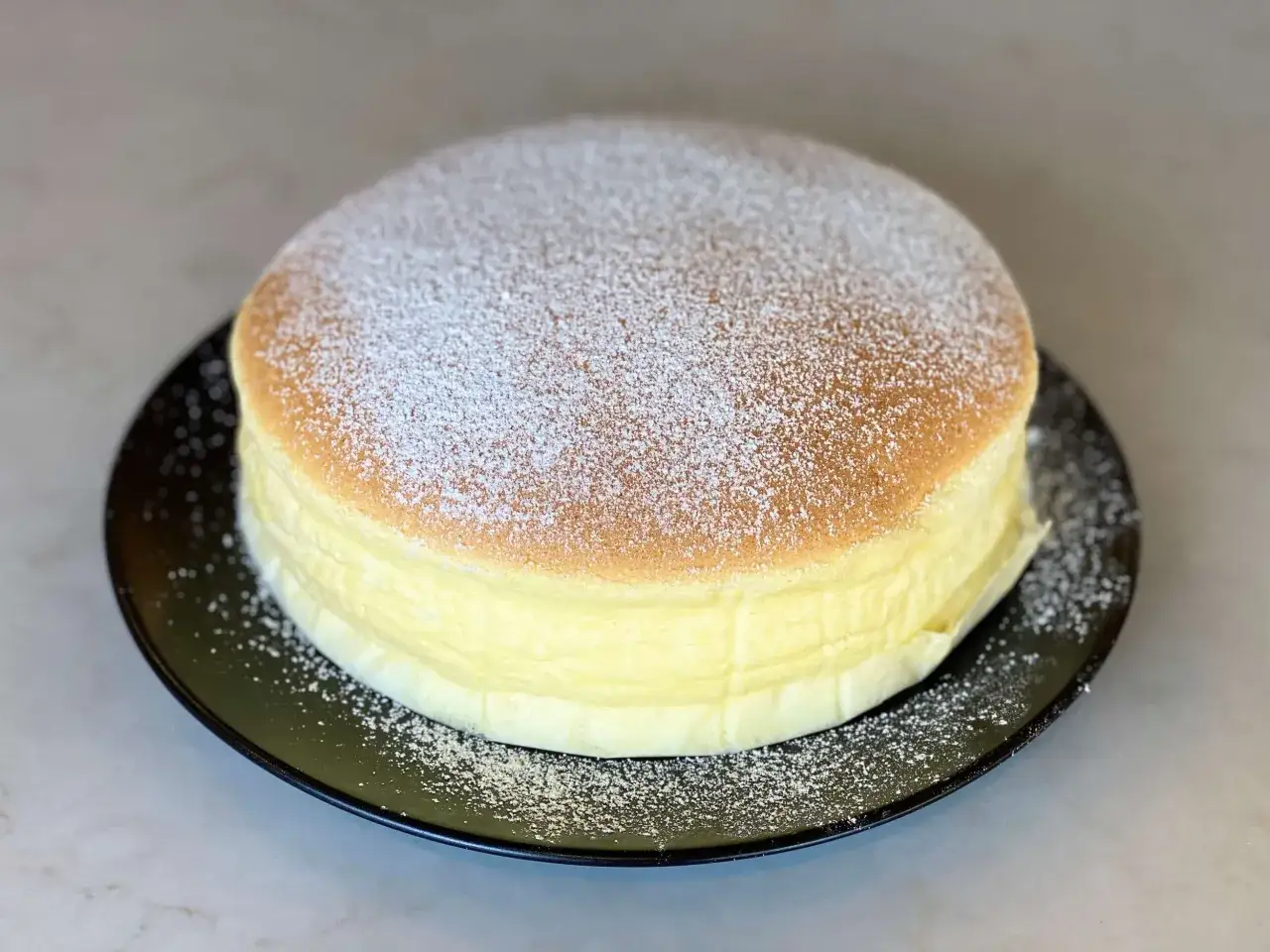 Un soffice cheesecake giapponese, leggero e arioso, spolverato di zucchero a velo su un piatto nero.