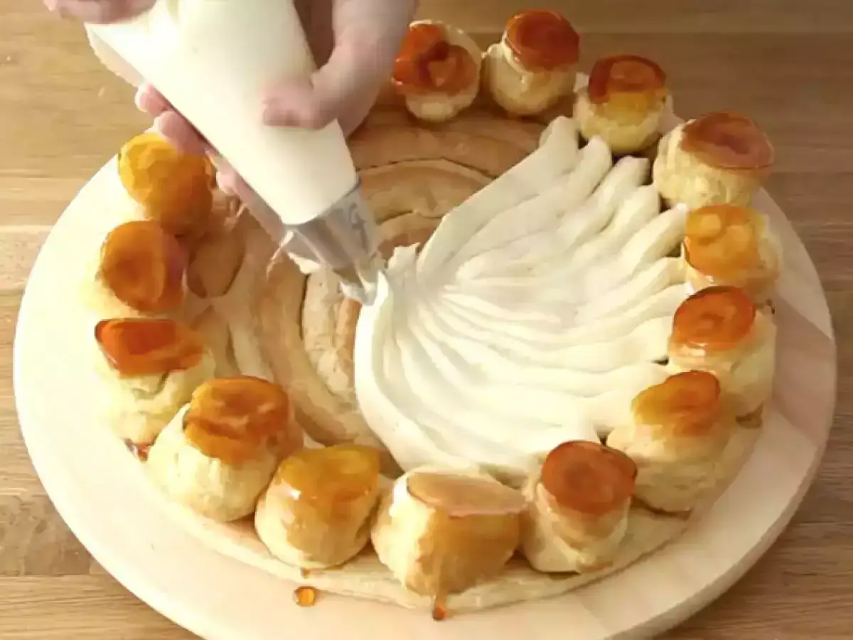 Una mano decora un dolce con crema chiboust, creando un effetto piumato. Piccoli bignè caramellati circondano la base.