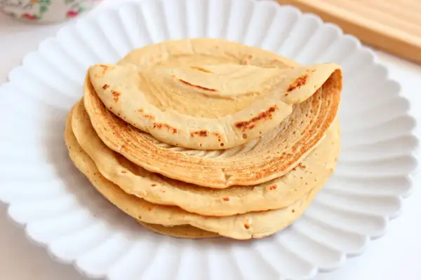 Pila di piadine di ceci dorate e leggermente maculate, servite su un piatto bianco a forma di fiore.