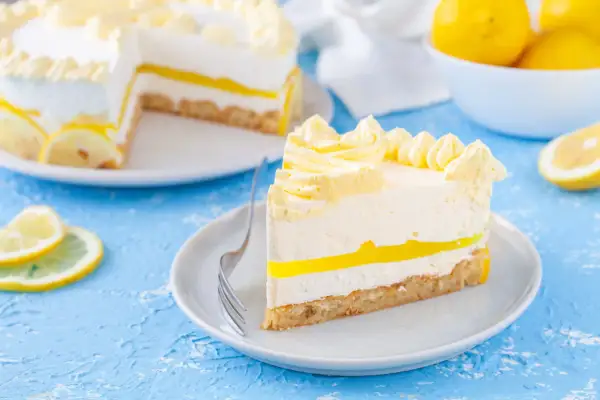 Una fetta di torta fredda al limone, con strati cremosi e una gelatina gialla brillante, decorata con ciuffi di panna e fette di limone.
