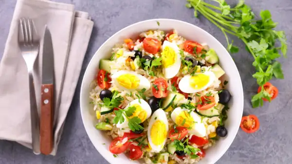 Una gustosa insalata di riso con uova sode, pomodorini, olive, mais e cetrioli, guarnita con prezzemolo fresco.