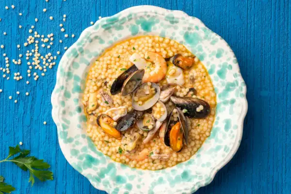 Piatto di fregola con pesce, cozze, vongole e gamberi, guarnito con prezzemolo tritato.