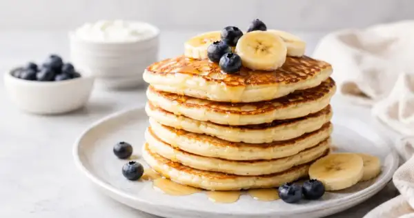 Pila di pancake proteici guarniti con mirtilli e banane, inzuppati nello sciroppo.