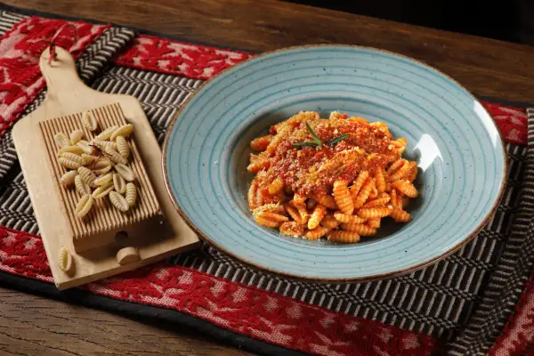 Gnocchetti sardi conditi con sugo rosso e parmigiano, accanto a una spianatoia con pasta fresca.