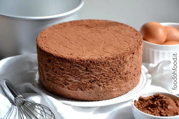 Un soffice pan di spagna al cioccolato, pronto per essere farcito. Accanto, uova fresche e cacao in polvere, ingredienti per un dolce perfetto.
