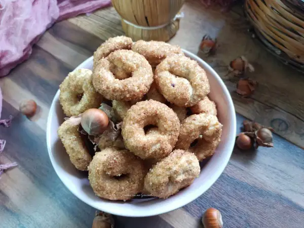 Ciambelline al vino cosparse di zucchero, con nocciole e un drappo rosa sullo sfondo.