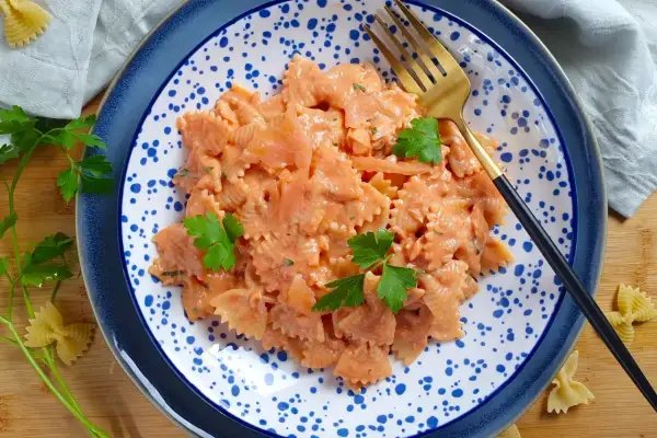 Farfalle al salmone cremose, guarnite con prezzemolo fresco, servite su un piatto blu e bianco con una forchetta dorata.