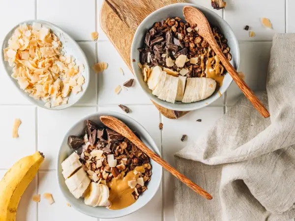 Due ciotole con granola fatta in casa, banana a fette, cioccolato fondente e burro d'arachidi. A lato, scaglie di cocco tostato.