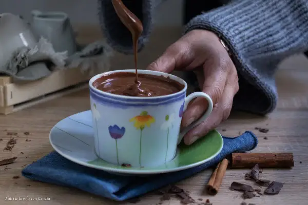 Una mano versa cioccolata calda in una tazza decorata con fiori. Cannella e scaglie di cioccolato completano la scena.