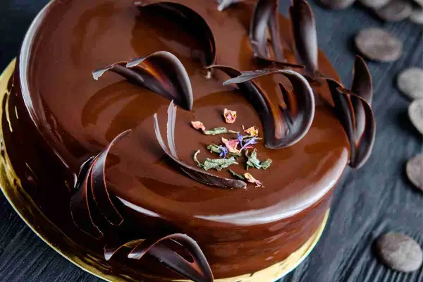 Una lucida torta Savoia al cioccolato, decorata con riccioli di cioccolato fondente e petali di fiori secchi.