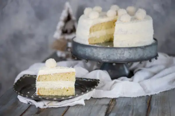 Una fetta di torta Raffaello, soffice e ricoperta di cocco, su un piatto decorato. Sullo sfondo, la torta intera su un'alzata.