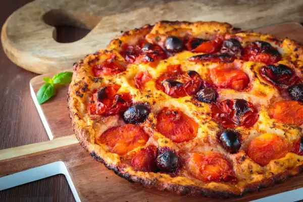 Focaccia pugliese dorata con pomodorini succosi e olive nere, guarnita con basilico fresco su tagliere di legno.