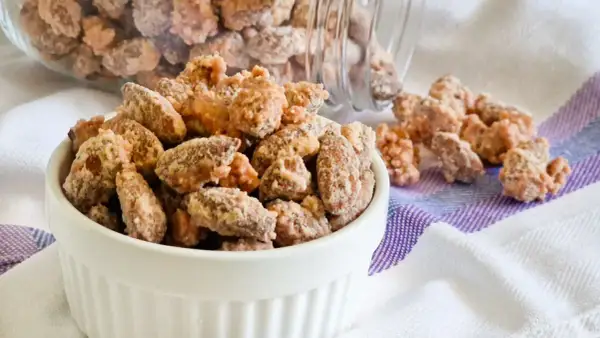 Un bianco contenitore pieno di mandorle pralinate, con altre sparse su un tovagliolo a righe.