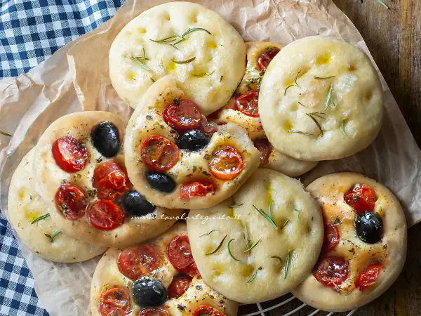 Focaccine in padella, alcune condite con pomodorini e olive nere, altre con rosmarino e olio.