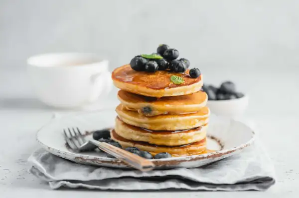 Pila di pancake con farina di riso, guarniti con mirtilli freschi e sciroppo d'acero, su un piatto rustico con una tazza di caffè sullo sfondo.