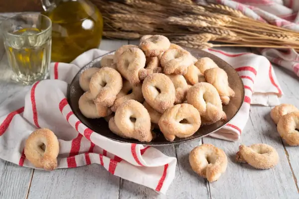 Un piatto colmo di taralli al vino bianco, cosparsi di zucchero, accanto a un bicchiere di vino e una bottiglia d'olio.