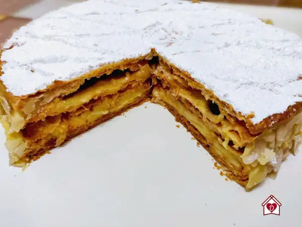 Una fetta di torta millefoglie con crema pasticcera e mandorle, cosparsa di zucchero a velo.