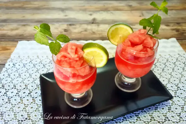 Due bicchieri di granita di anguria, guarniti con fettine di lime e foglie di menta, su un vassoio nero.