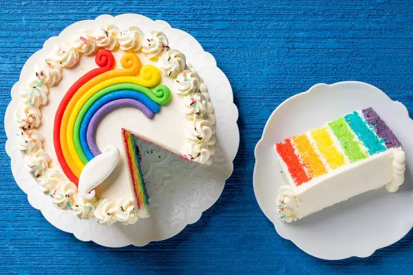 Torta arcobaleno con strati colorati e decorazioni di panna. Una fetta è già servita su un piattino.