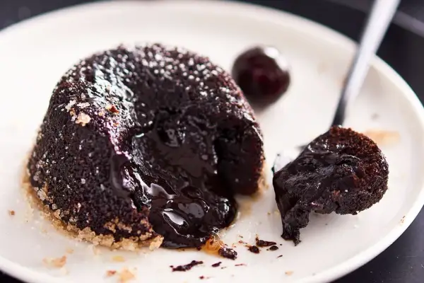 Un delizioso tortino al cioccolato con cuore fuso, cosparso di zucchero a velo, su un piatto bianco con una ciliegia e un cucchiaino.