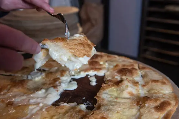 Una fetta di focaccia di Recco ricetta, filante e dorata, viene sollevata con una forchetta.
