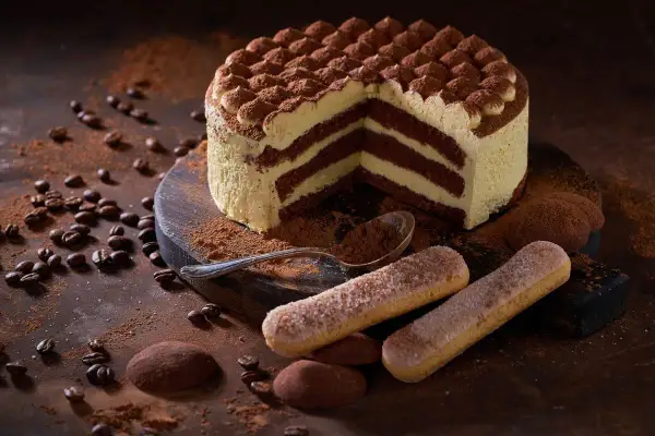 Una fetta di torta tiramisù, con strati di crema e cacao, circondata da savoiardi, chicchi di caffè e cacao in polvere.