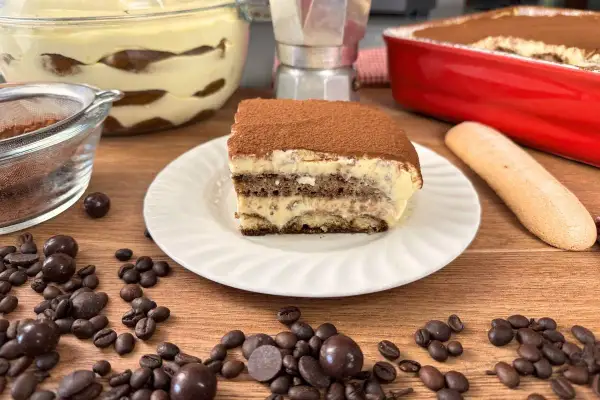Una fetta di tiramisù con uova pastorizzate, cosparso di cacao, su un piatto bianco. Intorno, chicchi di caffè, palline di cioccolato e un savoiardo.