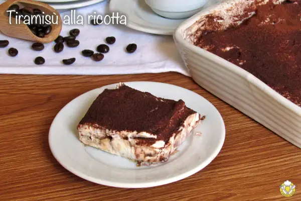 Una fetta di tiramisù alla ricotta, cosparso di cacao, su un piatto bianco, con chicchi di caffè e una teglia sullo sfondo.