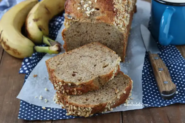Fetta di banana bread senza burro, soffice e profumato, con crosta croccante di frutta secca. Accanto, banane mature e una tazza blu.