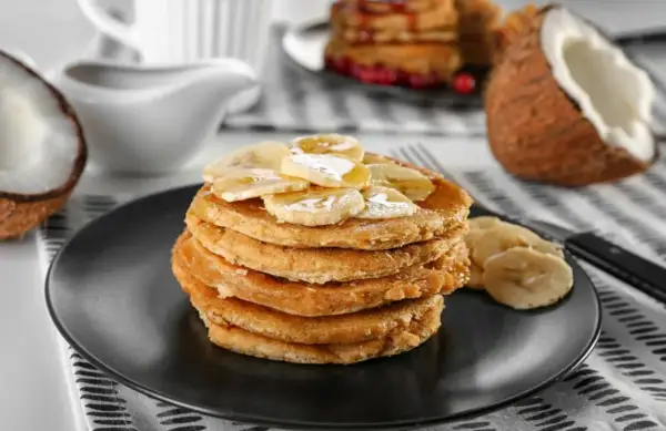 Pancakes con farina di cocco, guarniti con banane a fette e sciroppo, su un piatto nero. A lato, un cocco aperto.